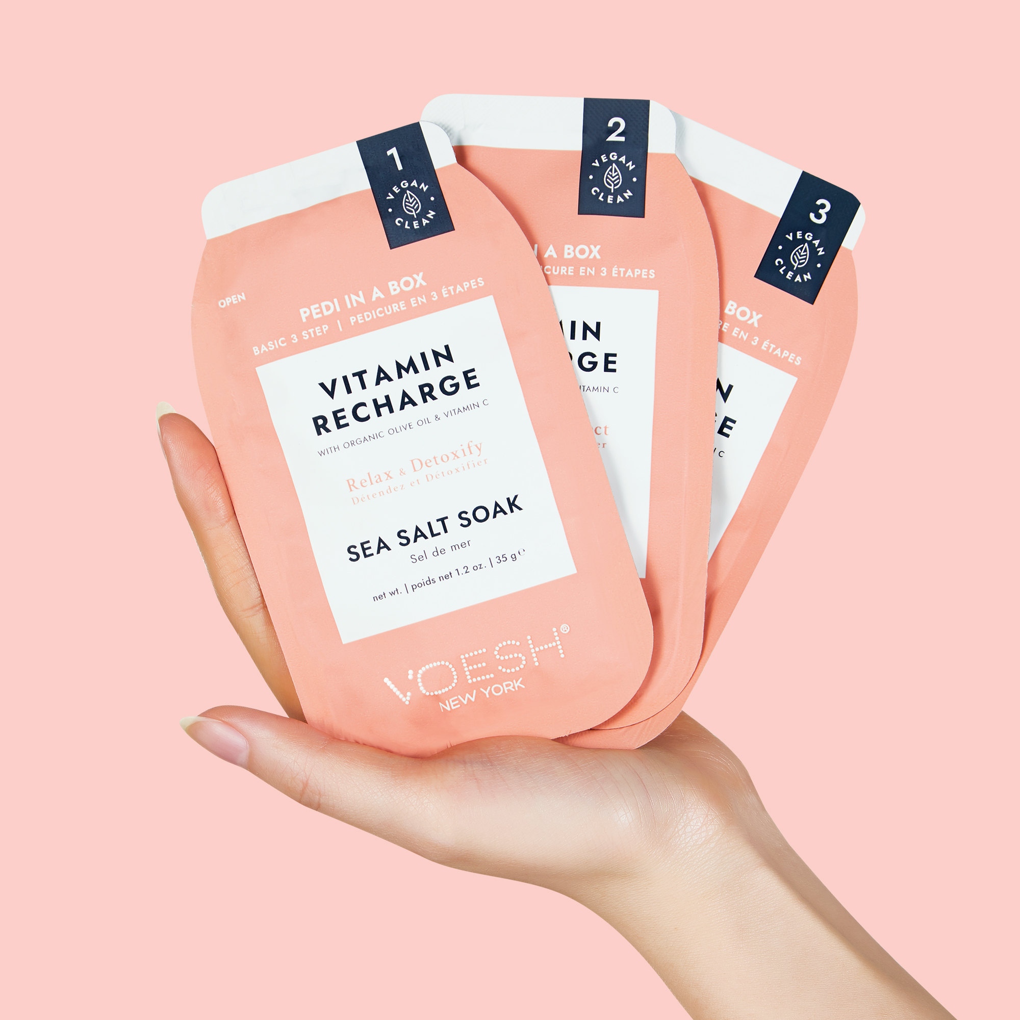 Hand hält VOESH New York Pedi in a Box Vitamin Recharge, ein veganes 3-Schritt-Pediküre-Set mit Sea Salt Soak, auf rosa Hintergrund