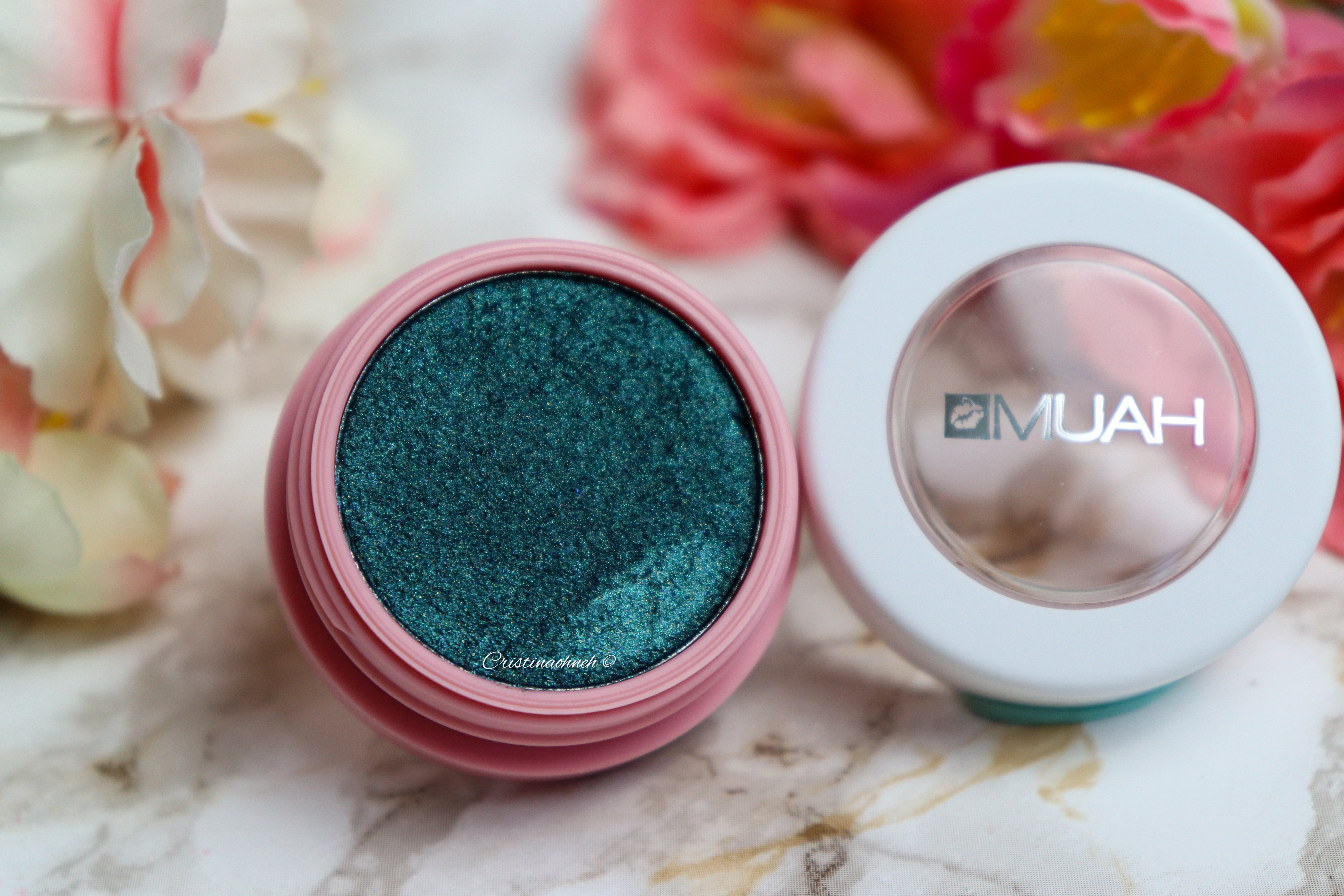 Döschen mit schimmerndem türkisfarbenem Lidschatten von MUAH, umgeben von Blüten.