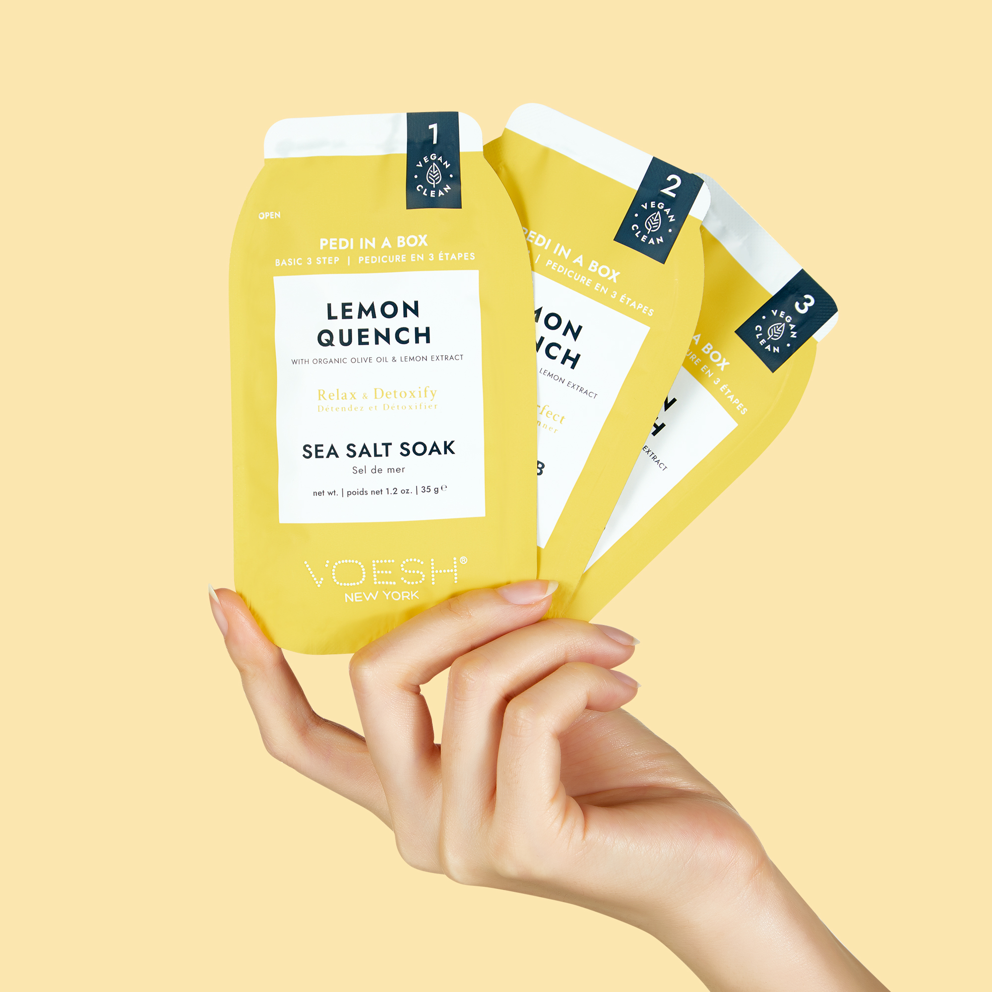Hand hält VOESH New York Pedi in a Box Lemon Quench, ein 3-Schritt-Pediküre-Set mit Sea Salt Soak, Scrub und Massage Butter in gelben Sachets