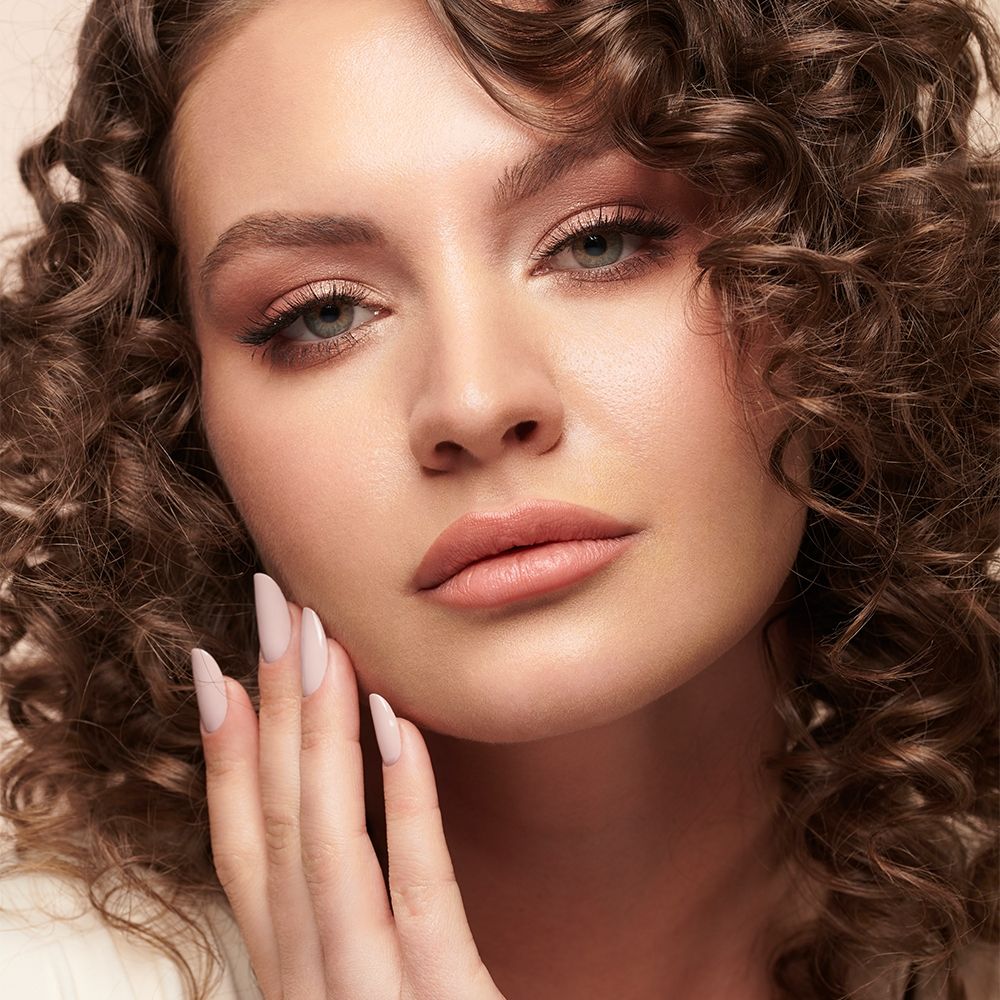 Nahaufnahme einer Frau mit lockigem Haar, natürlichem Make-up und nudefarbenen, mandelförmigen Fingernägeln als Produktdarstellung für Nageldesign oder Kunstnägel