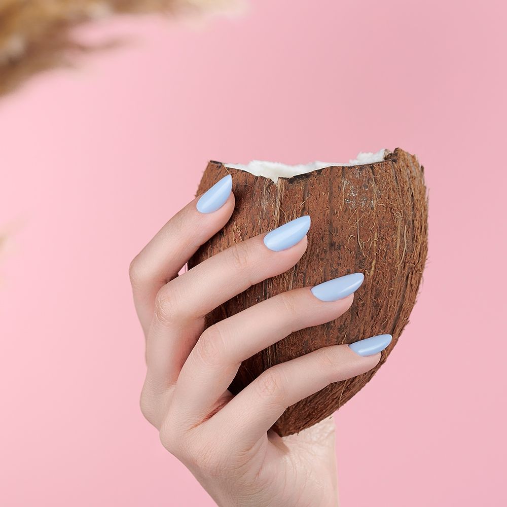 Nahaufnahme einer Hand mit hellblauen, mandelförmigen Nägeln, die eine halbe Kokosnuss vor rosa Hintergrund hält – Produktdarstellung für Nagellack oder künstliche Nägel