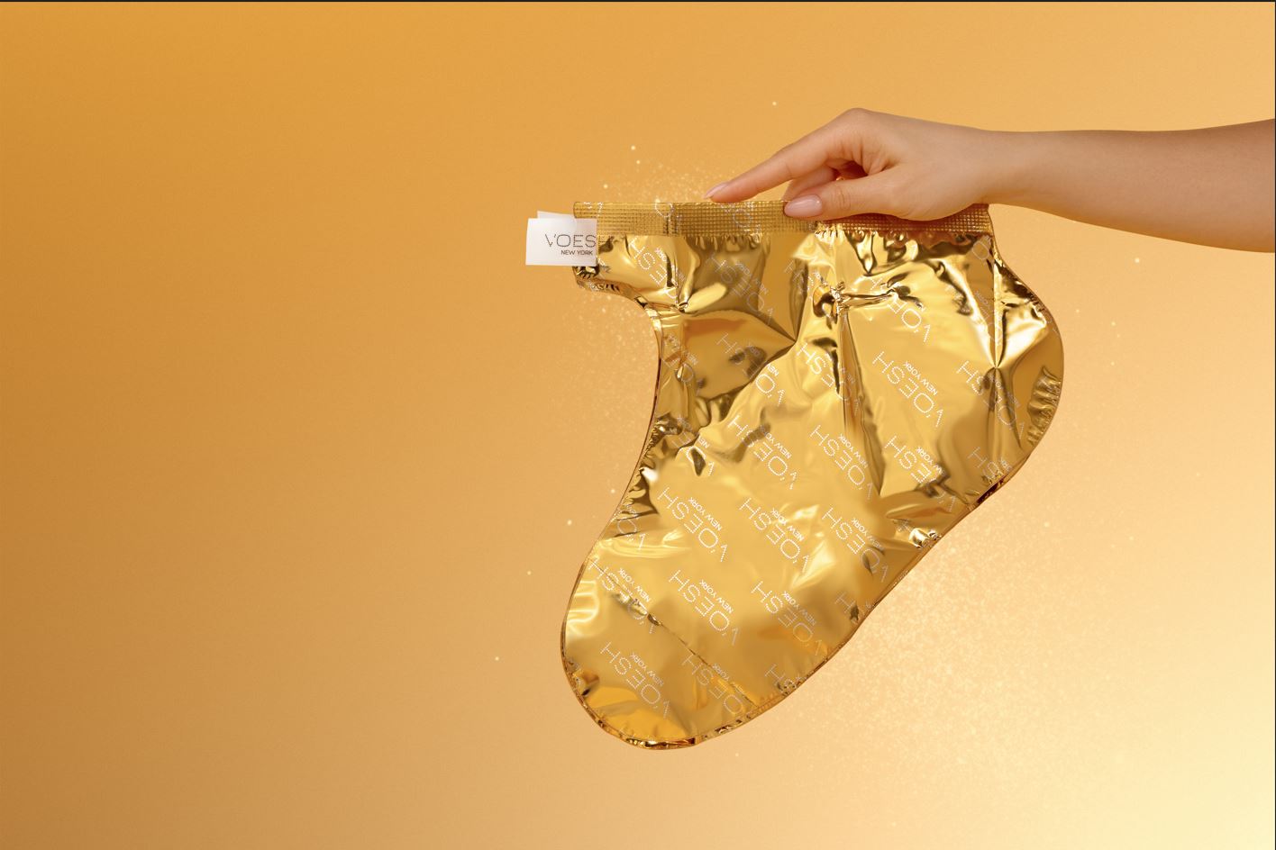 Goldene Fußmaske in Sockenform von VOESH New York, gehalten von einer Hand vor warm goldenem Hintergrund