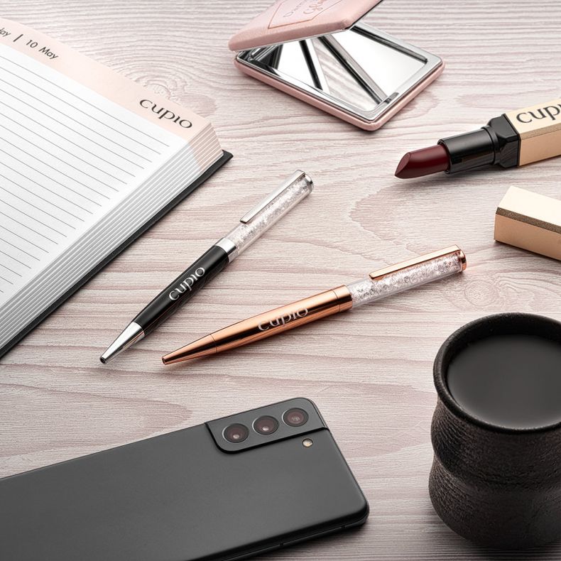 Schreibtisch-Setup mit Notizbuch, zwei Stiften, Lippenstift, Handspiegel, Smartphone und einer Tasse