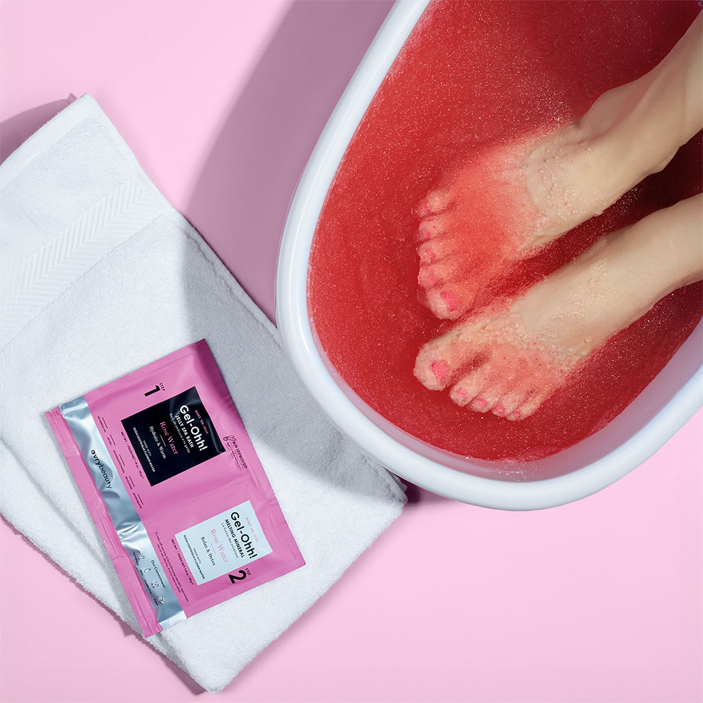 Füße in einer Schüssel mit rosa Gel-Ohh! Fußbad, daneben zwei Gel-Ohh! Pflegebeutel auf weißem Handtuch