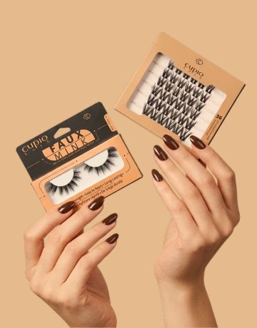 Zwei Hände mit dunkel lackierten Nägeln präsentieren Cupio Faux Mink künstliche Wimpern und Wimpernbüschel in beige-schwarzer Verpackung.
