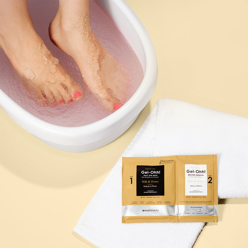 Füße in rosa Fußbad neben Gel-Ohh! Fußpflege-Set von avrybeauty auf weißem Handtuch für Pediküre und Pflege zuhause
