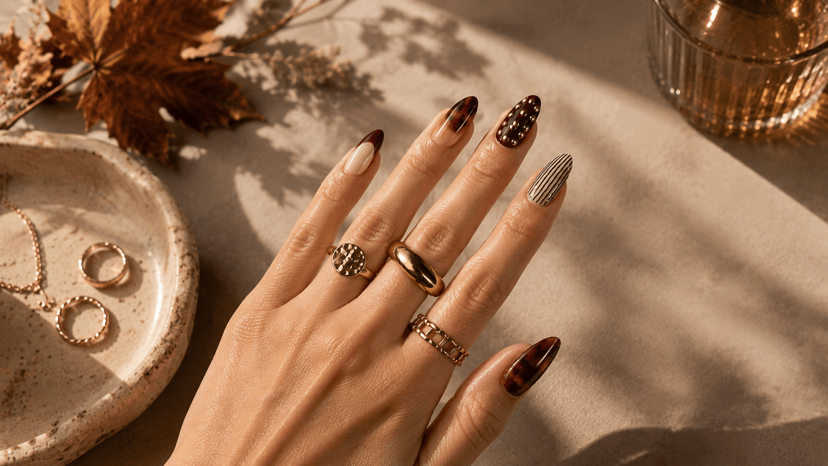 Nahaufnahme einer Hand mit langen mandelförmigen Fingernägeln im herbstlichen Design in Braun, Creme, Leo-Optik, Nieten und Streifen, kombiniert mit goldfarbenen Ringen
