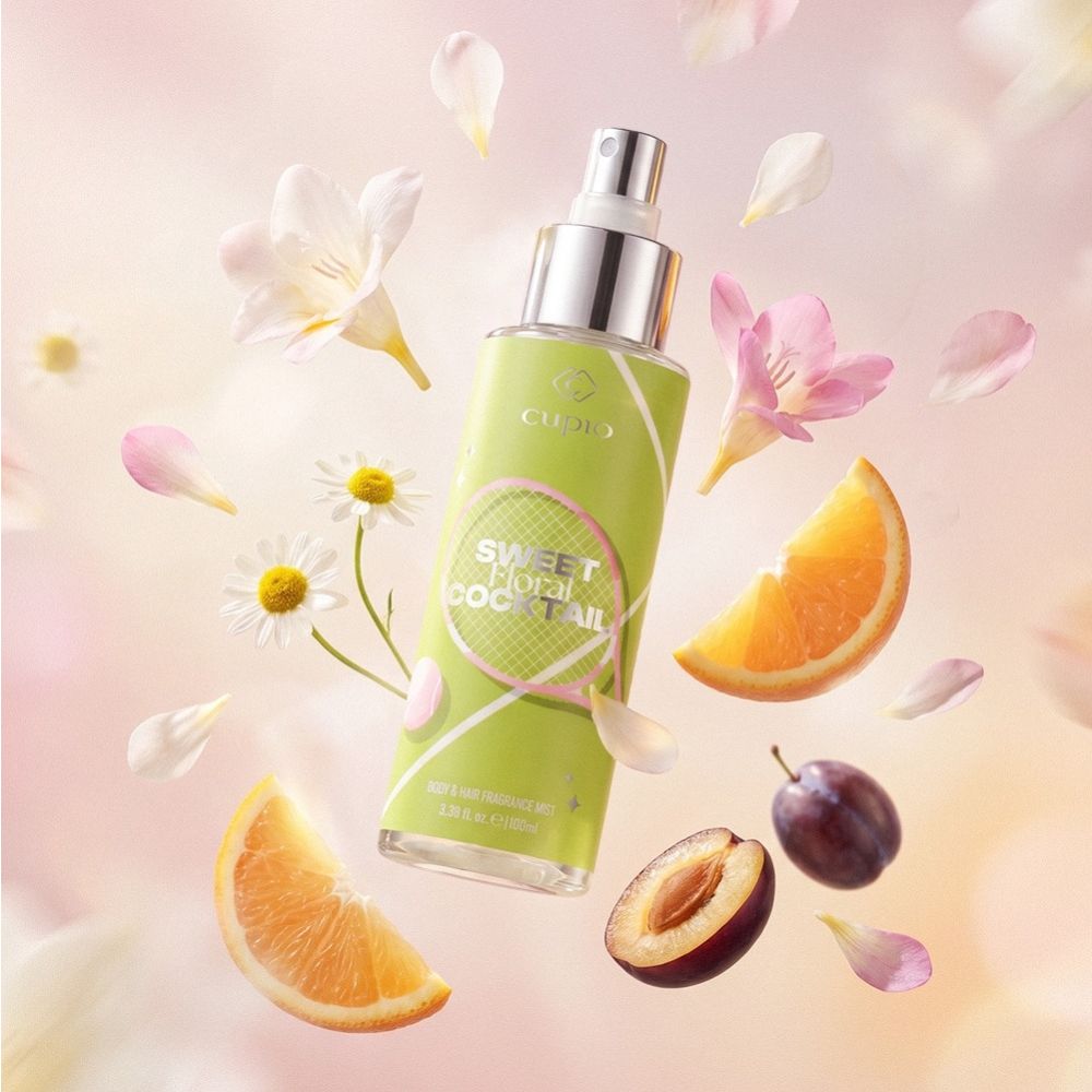 Cupio Sweet Fruit Cocktail Body & Hair Fragrance Mist, grüner Sprayflakon mit Orange, Pflaume, Kamille und Blütenblättern vor zartem Hintergrund