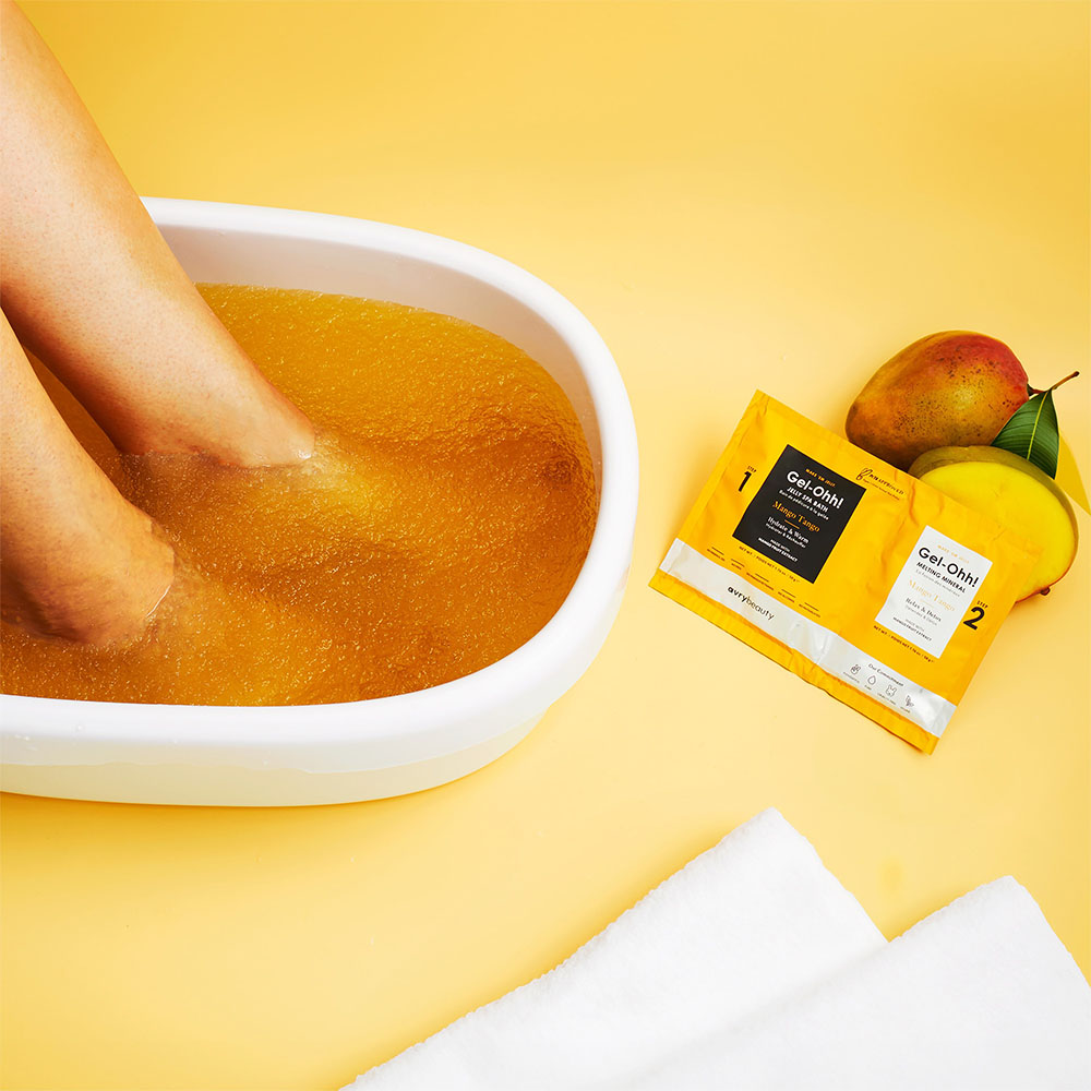 Füße in einer Schale mit orangefarbenem Gelbad neben GelOhh! Mango Fußmasken-Beuteln und frischer Mango