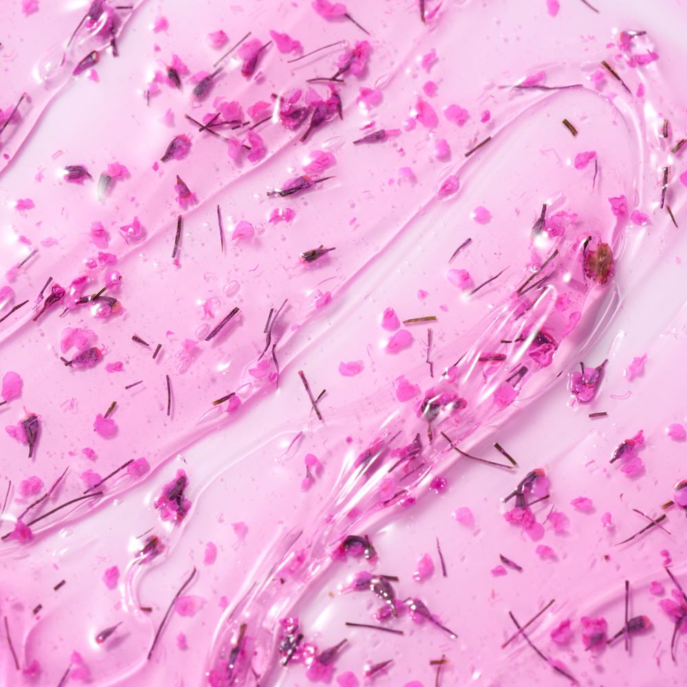 Nahaufnahme einer rosa Gel-Textur mit feinen Blüten- und Kräuterpartikeln als Produktbild für ein pflegendes Kosmetikprodukt