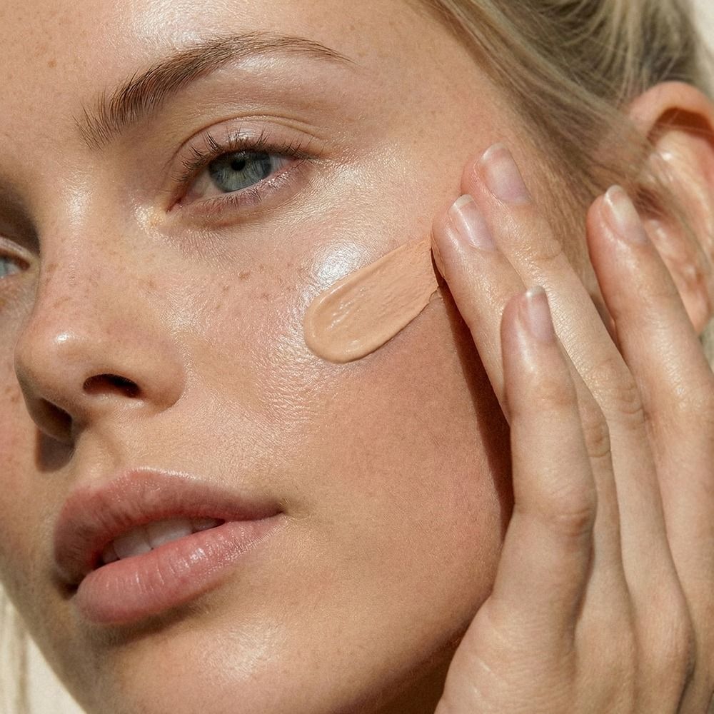 Nahaufnahme eines Gesichts, auf dessen Wange mit den Fingern eine flüssige Foundation für ein natürlich strahlendes Make-up aufgetragen wird