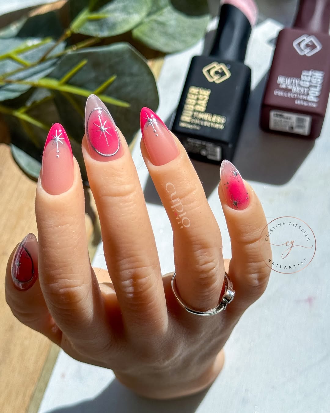Detailaufnahme einer Hand mit elegantem Nageldesign in Pink und klaren Farben, umgeben von Nagellackflaschen.