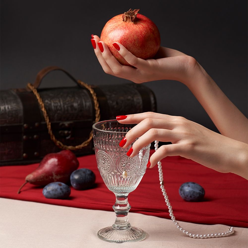 Hand mit Granatapfel über einem kunstvollen Glas, umgeben von Früchten und einer Schatztruhe