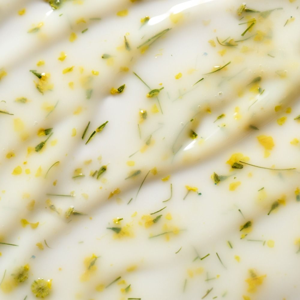 Nahaufnahme einer weißen Creme mit feinen Kräutern und gelben Blütenpartikeln als Produkttextur für Naturkosmetik