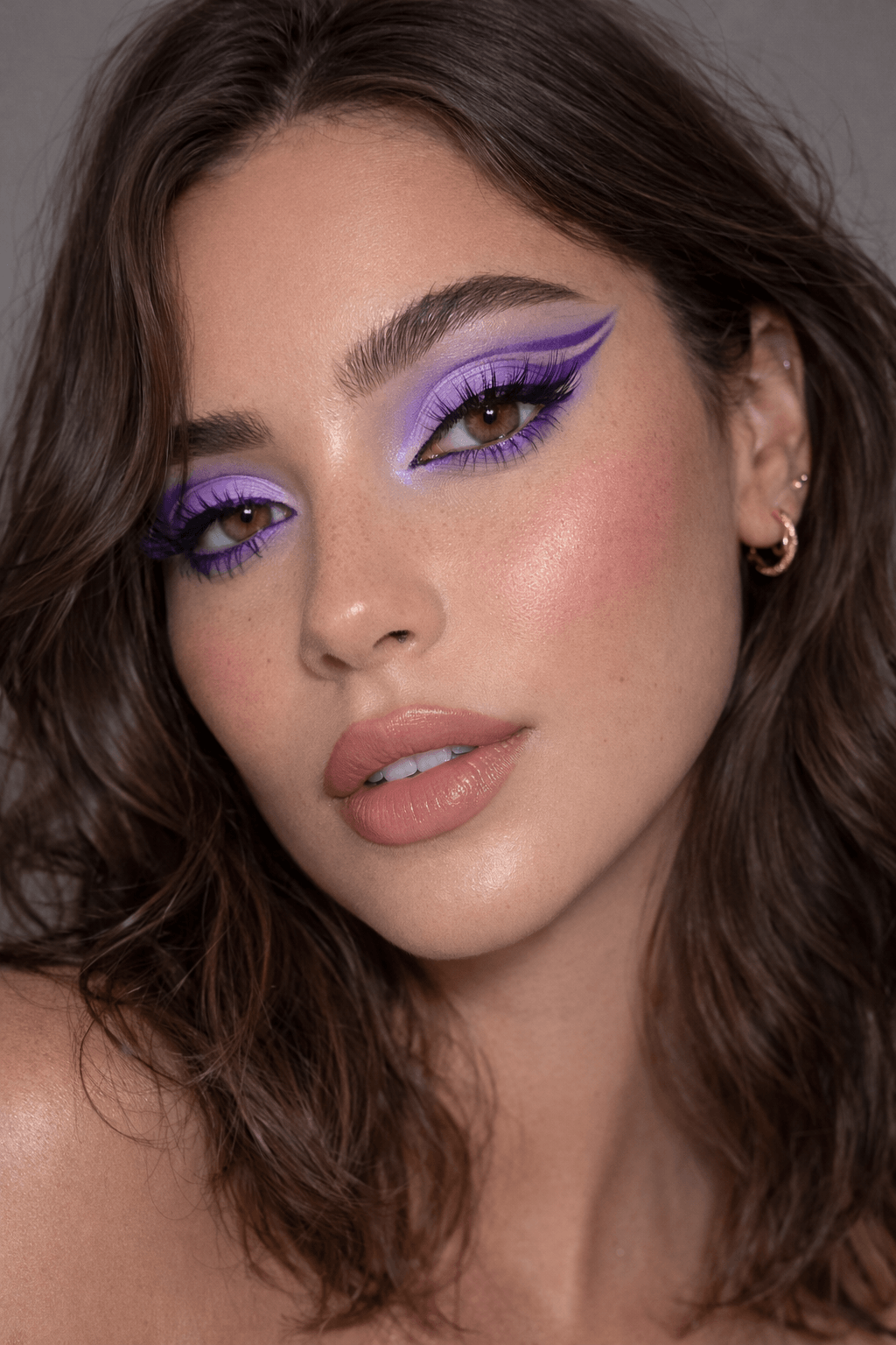 Nahaufnahme eines Gesichts mit auffälligem lila Lidschatten, Graphic Eyeliner, langen Wimpern und natürlichen Nude-Lippen