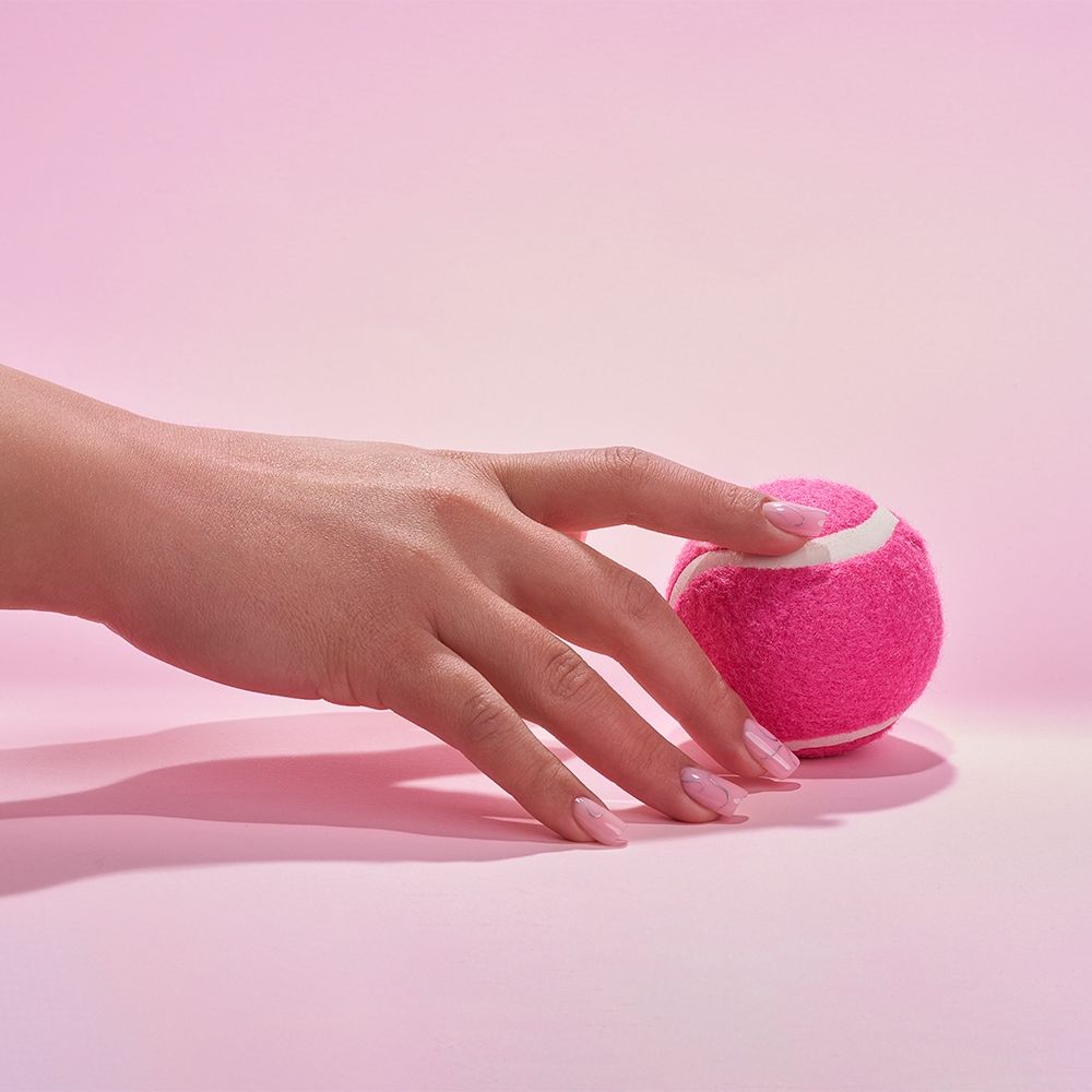 Nahaufnahme einer Hand, die einen pinken Tennisball auf einem rosa Hintergrund berührt.