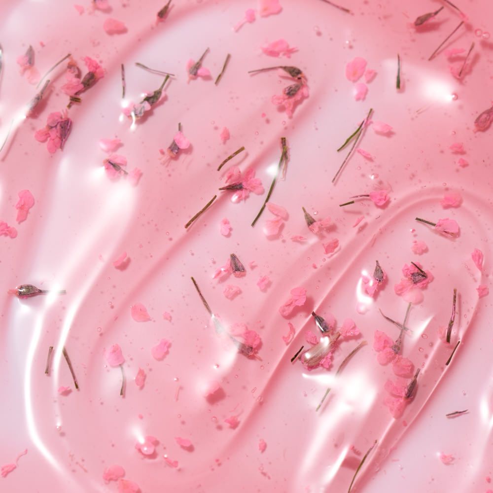 Rosafarbene, glänzende Gel- oder Serum-Textur mit feinen Blütenblättern und Pflanzenfasern als Produktbild für Naturkosmetik oder Hautpflege