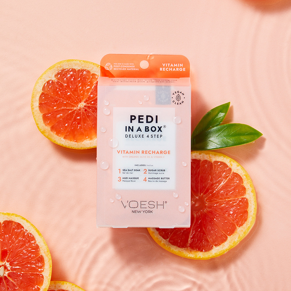 Voesh Pedi in a Box Deluxe 4 Step Vitamin Recharge, 4-Stufen-Spa-Pediküre mit Bio-Olivenöl und Vitamin C, Verpackung zwischen Grapefruithälften auf rosafarbenem Hintergrund