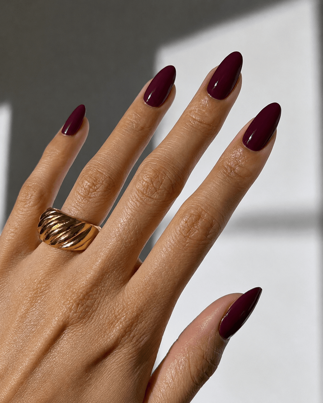 Nahaufnahme einer Hand mit dunkelrot lackierten, mandelförmigen Nägeln und einem breiten goldfarbenen Statement-Ring