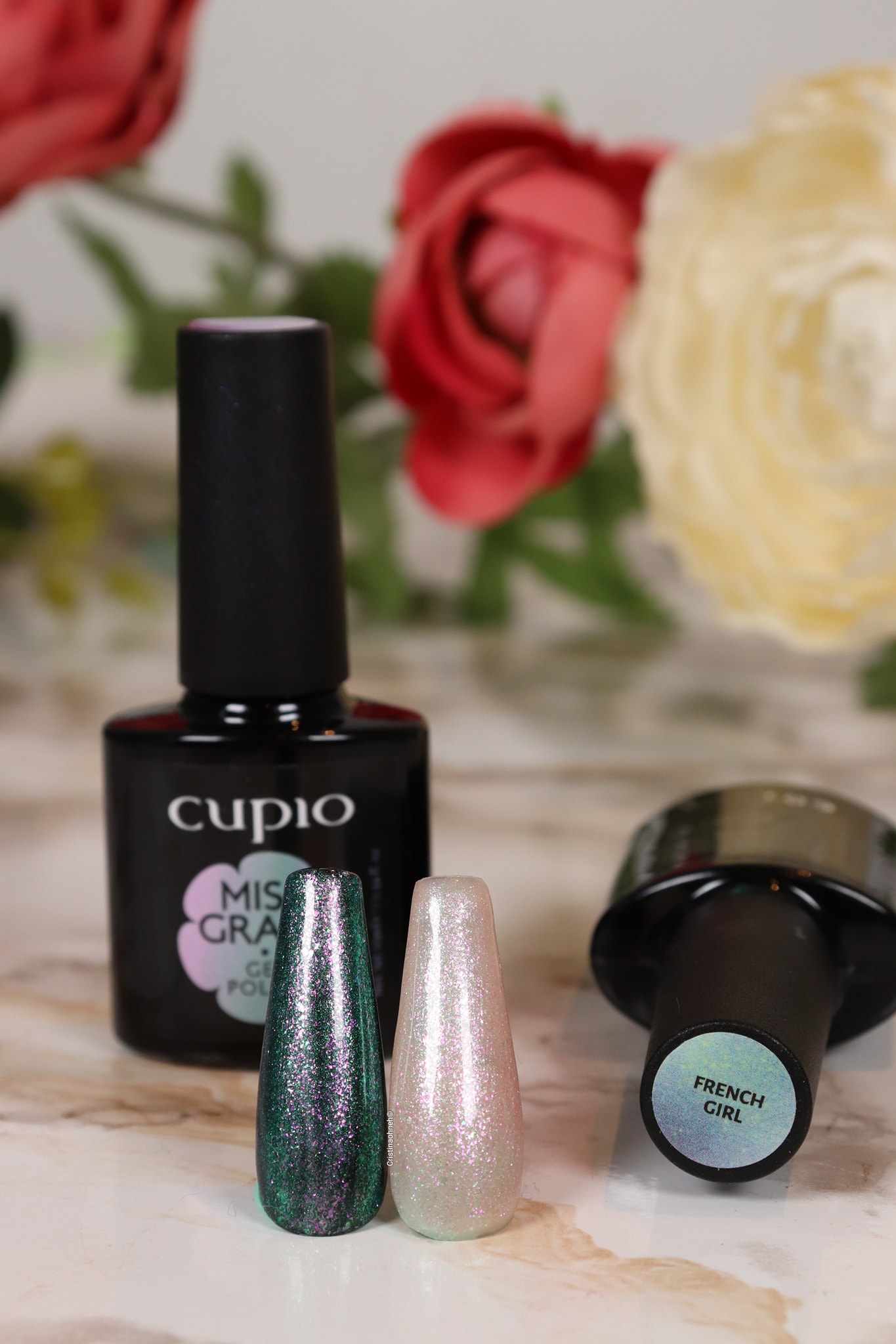 273940472_10221077892199744_8466062528911026222_n Zwei farbige Nagellacke von Cupio neben einer Flasche, dekoriert mit Blumen im Hintergrund.