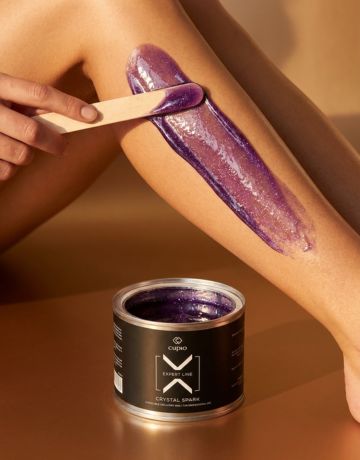 Nahaufnahme: Lila Crystal Shine Enthaarungswachs wird mit einem Holzspatel auf das Bein aufgetragen, der Wachstiegel steht davor.