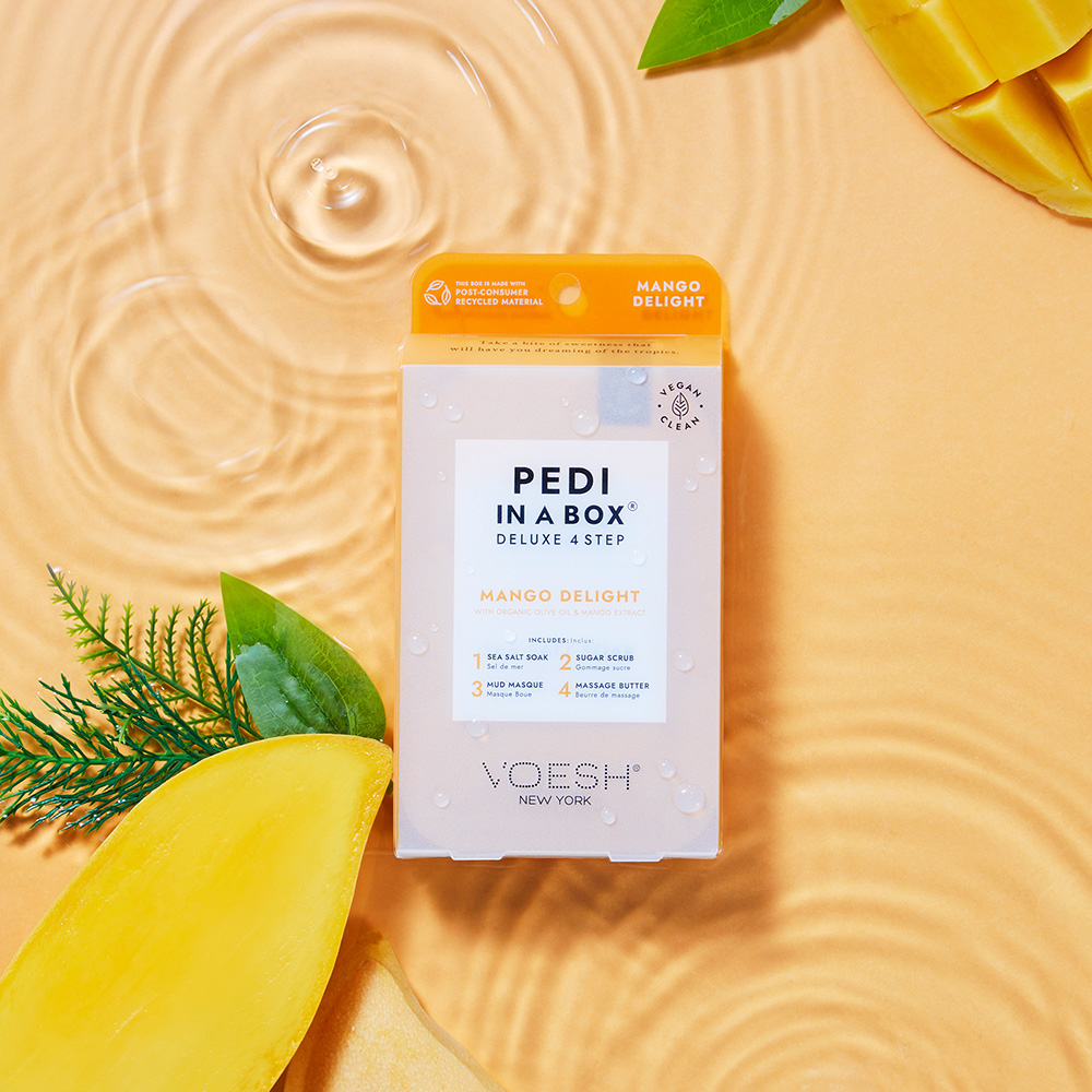 VOESH Pedi in a Box Deluxe 4 Step Mango Delight Pediküre-Set, umgeben von frischer Mango und Blättern auf goldenem Wasserhintergrund