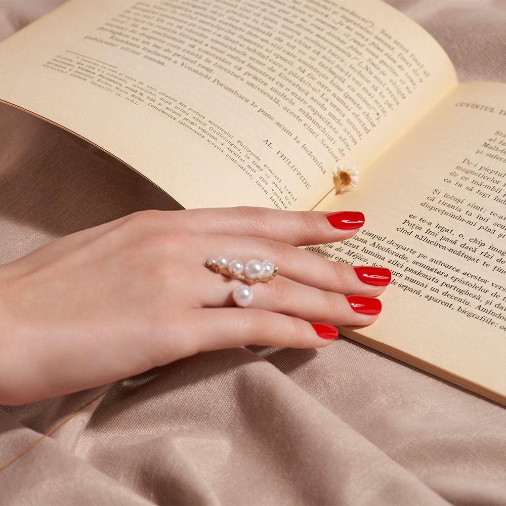 Hand mit einem eleganten Ring aus Perlen und rotem Nagellack vor einem offenem Buch