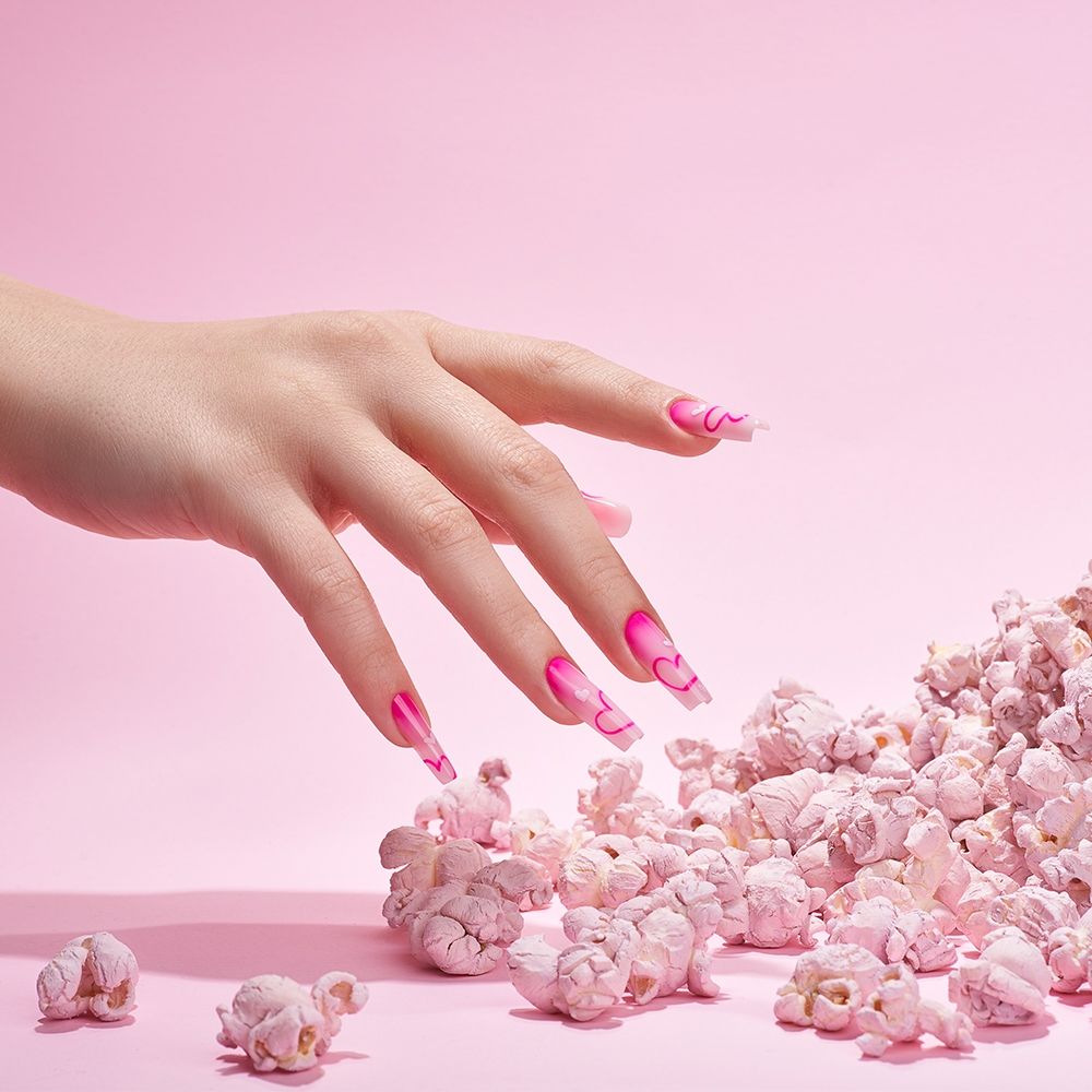 Ein Hand mit auffälligem pinken Nageldesign, umgeben von pinkem Popcorn auf einem rosa Hintergrund.