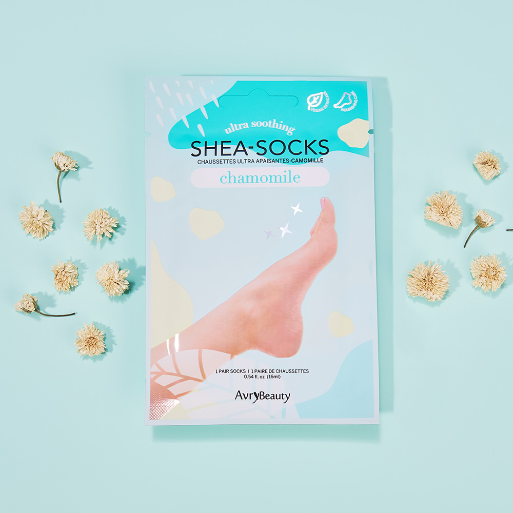 Verpackung der AvryBeauty Shea-Socks Chamomile, ultra beruhigende Fußmaske mit Shea-Butter, auf hellblauem Hintergrund mit getrockneten Kamillenblüten