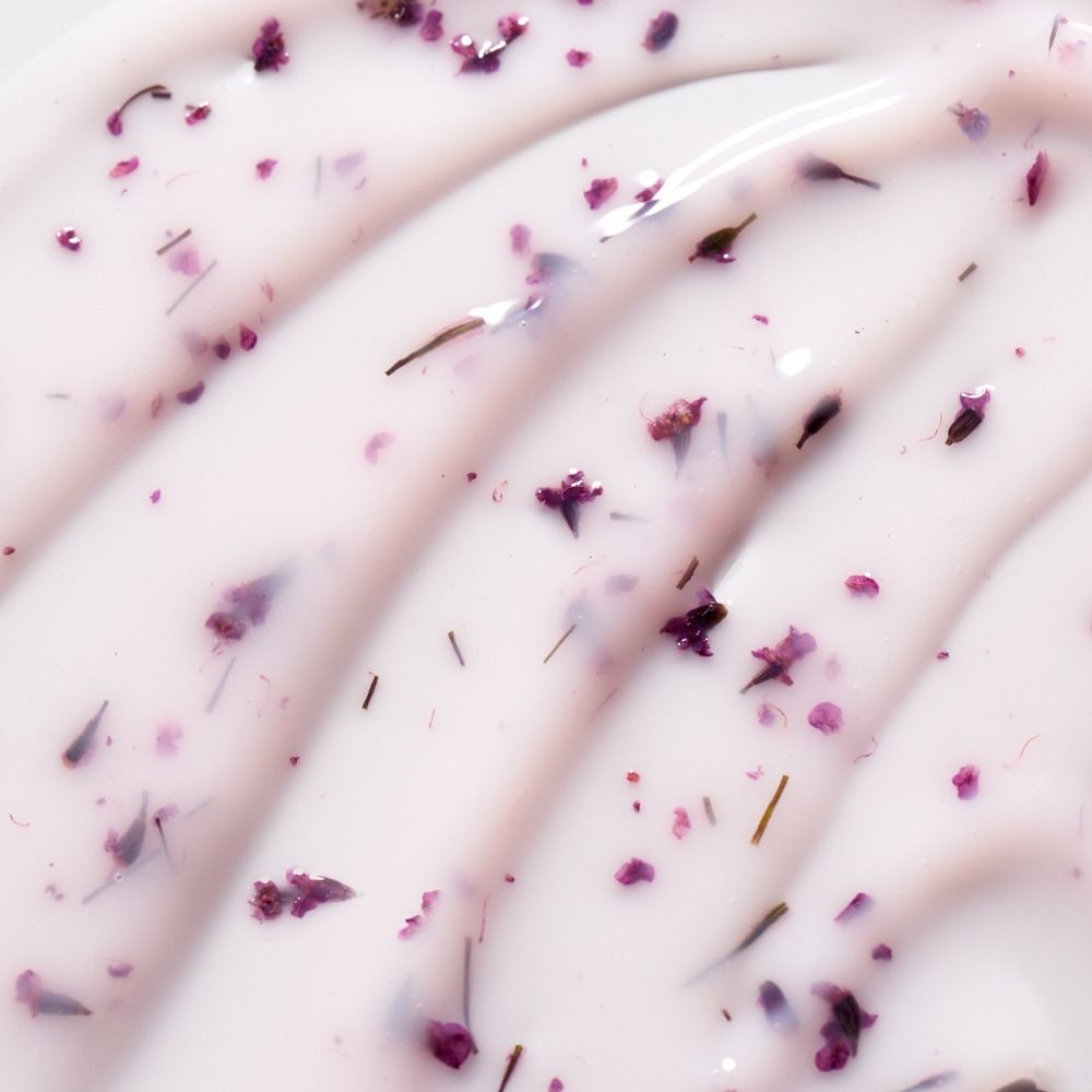 Nahaufnahme einer weißen Pflegecreme mit eingearbeiteten violetten Blüten- und Kräuterpartikeln, cremige Produkttextur im Makro